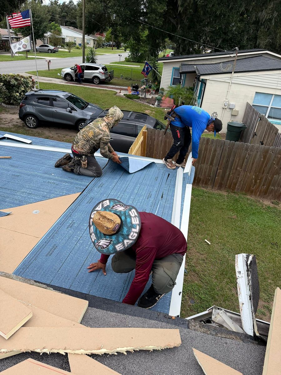 Three roofers laying blue underlayment on a home while the old roof edge is torn off